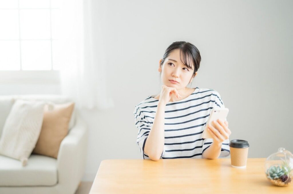 スマホを持ちながら机に肘を置いて考えるボーダーシャツの女性